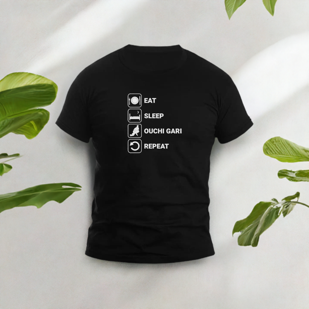 Black judo-themed t-shirt with 'Eat Sleep Ouchi Gari Repeat' text and icons, displayed with green leaves for a fresh aesthetic. Unisex design.