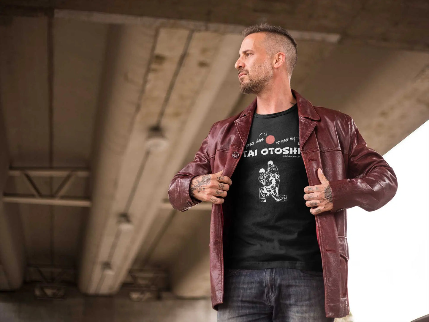 Man wearing a black 'Tai Otoshi Press Here' Judo T-Shirt styled with a red leather jacket, standing under a bridge.