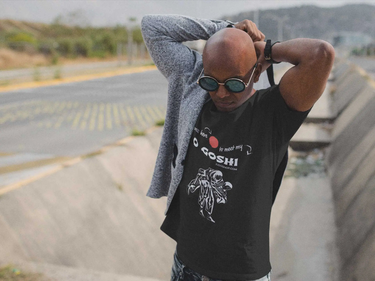Man adjusting his sweater outdoors, showcasing a black Judo T-Shirt with 'Press Here to Meet My O Goshi' text and martial arts graphic.