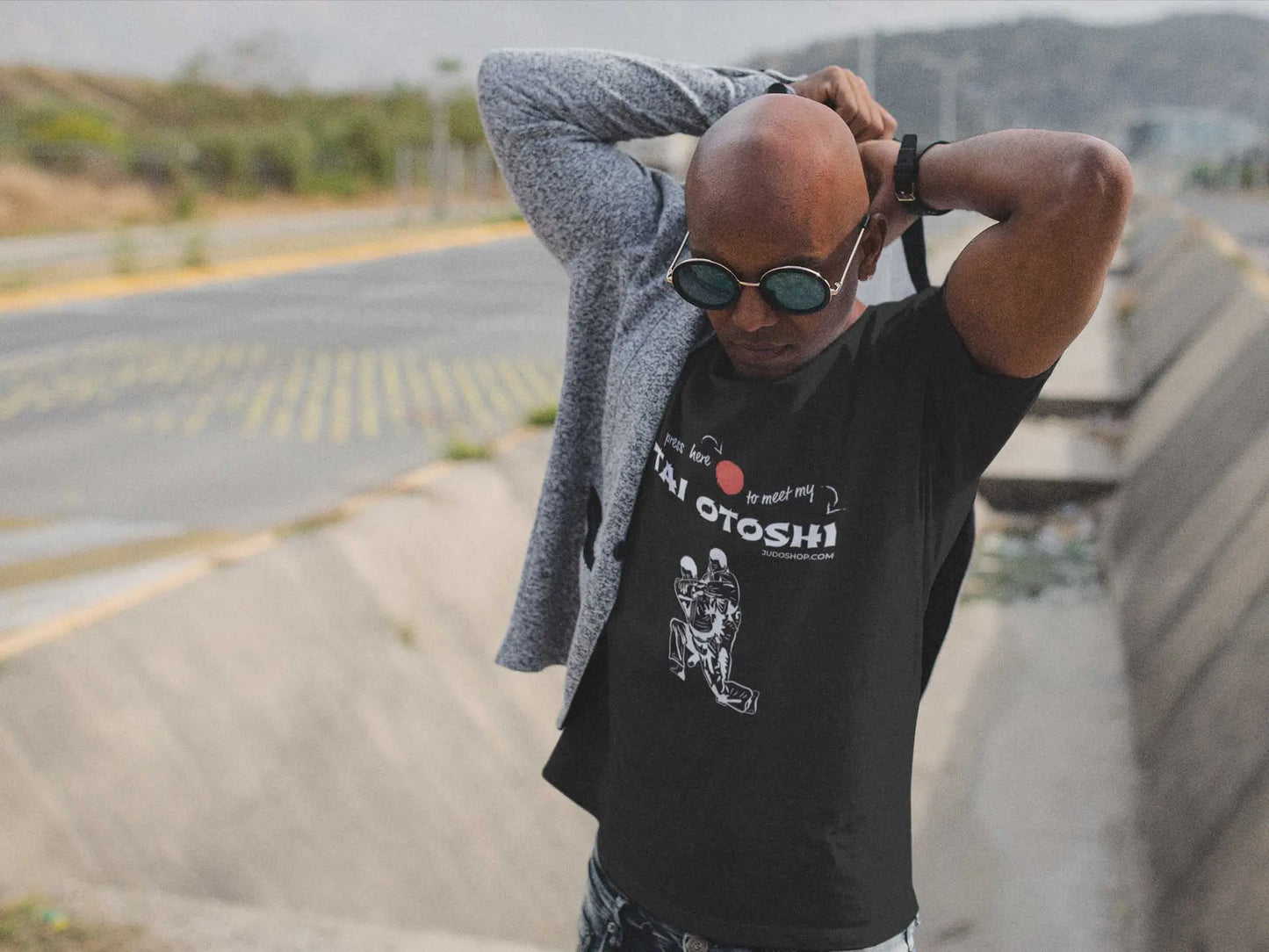 Man adjusting his jacket while wearing a black 'Tai Otoshi Press Here' Judo T-Shirt, standing near a road.
