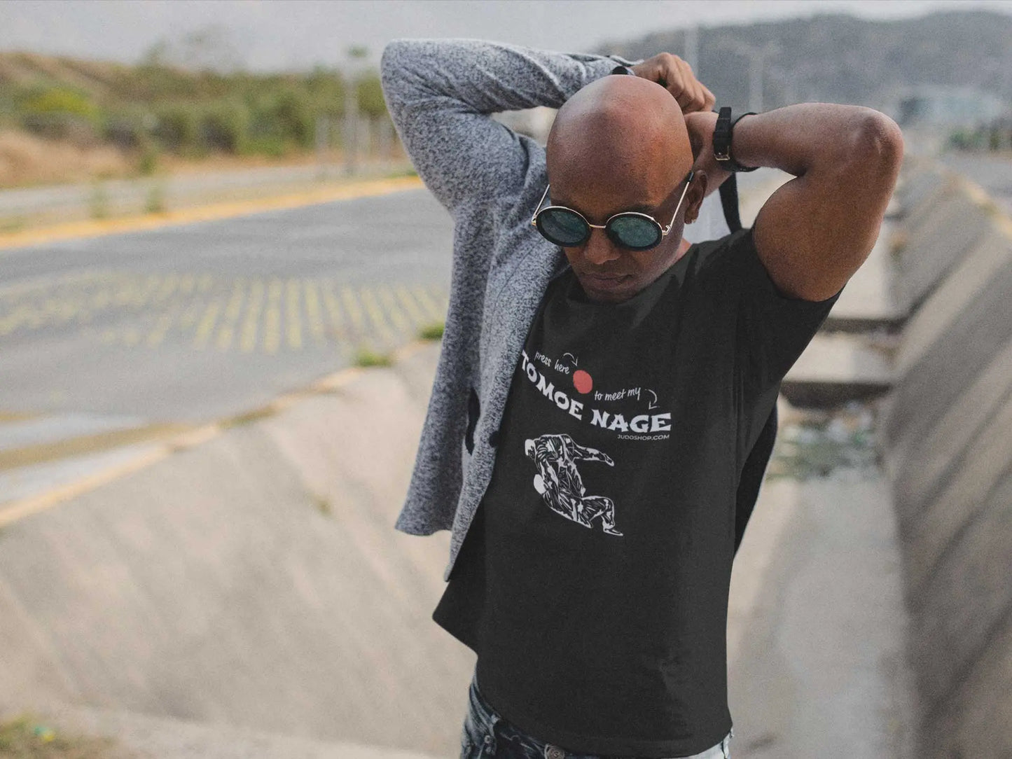 Man adjusting his jacket while wearing a black Judo T-Shirt with 'Press Here to Meet My Tomoe Nage' design, styled with sunglasses for a modern look.