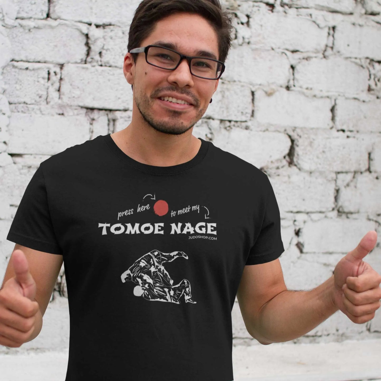 Smiling man giving thumbs up while wearing a black Judo T-Shirt with 'Press Here to Meet My Tomoe Nage' design, styled for casual wear.