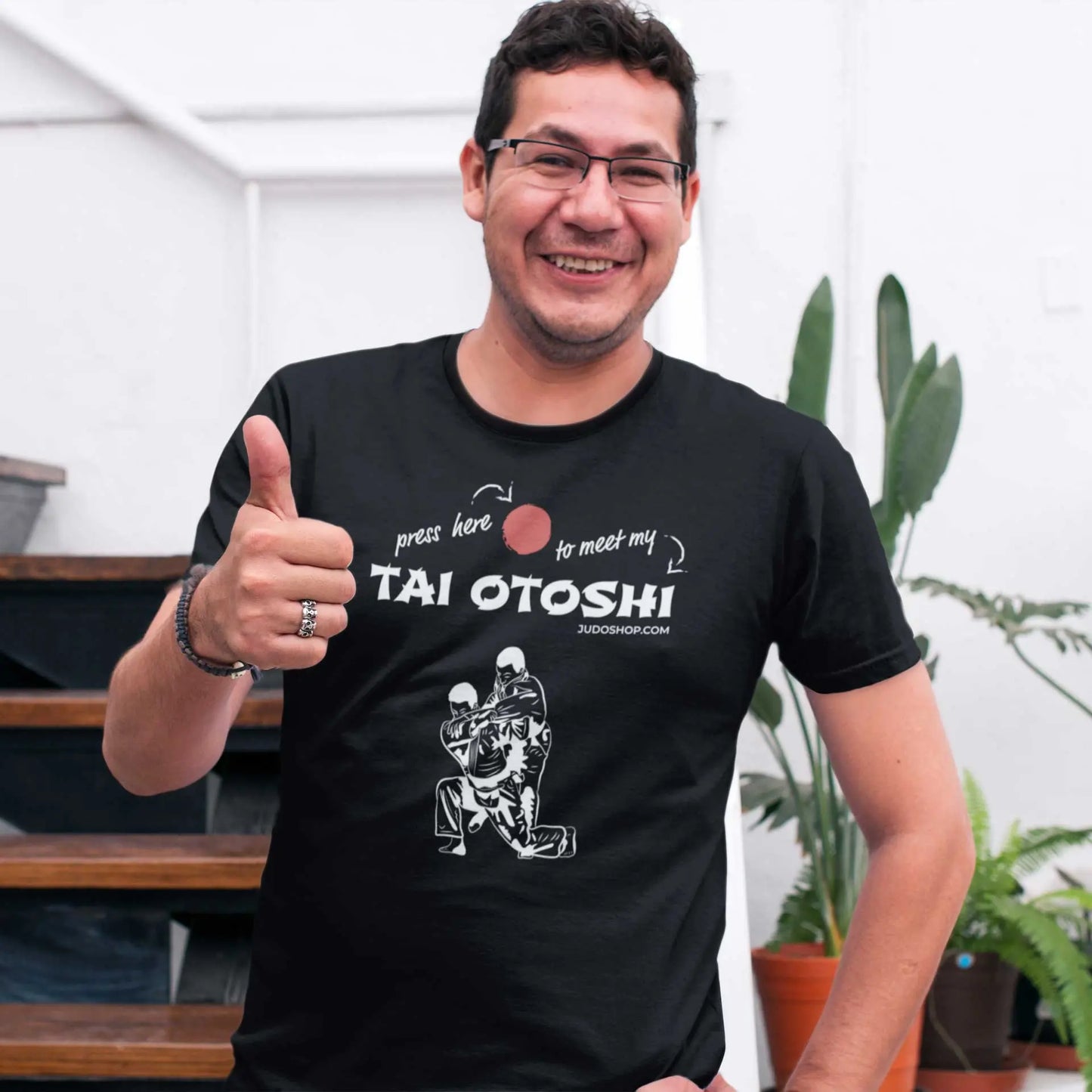 Smiling man giving a thumbs-up while wearing a black 'Tai Otoshi Press Here' Judo T-Shirt, standing indoors.