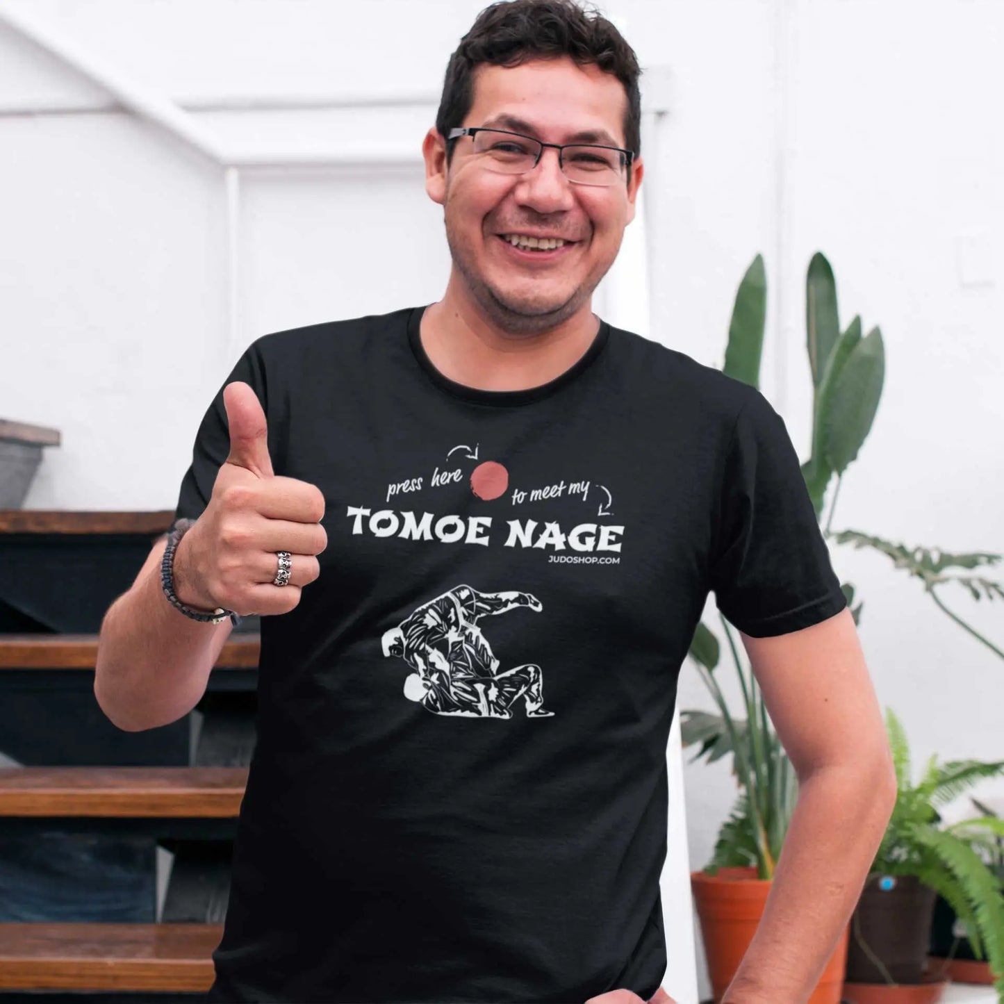 Man posing indoors in a black Judo T-Shirt with 'Press Here to Meet My Tomoe Nage' design, paired with glasses and casual accessories.