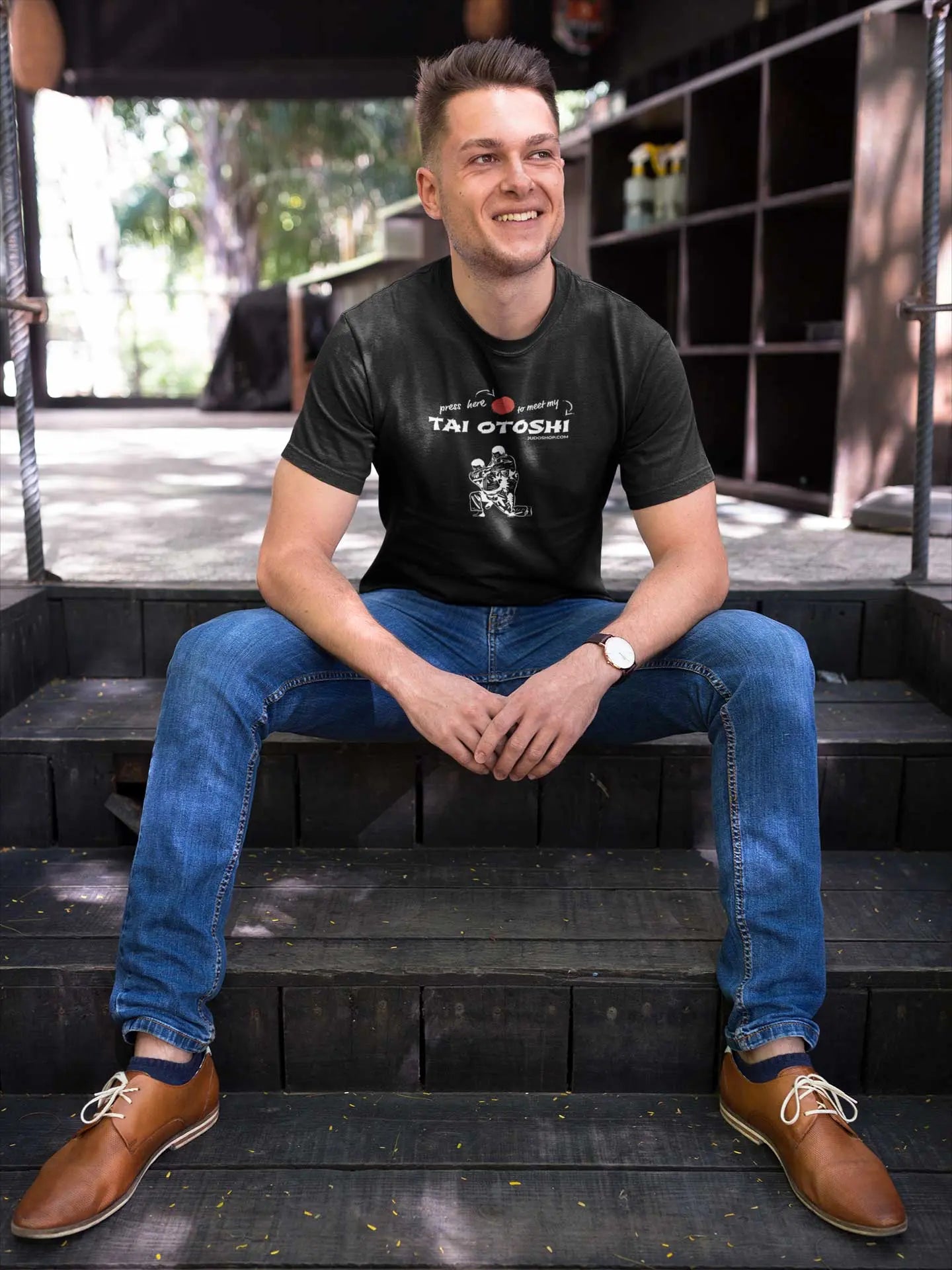 Man sitting on wooden steps wearing a black 'Tai Otoshi Press Here' Judo T-Shirt paired with blue jeans and brown shoes.