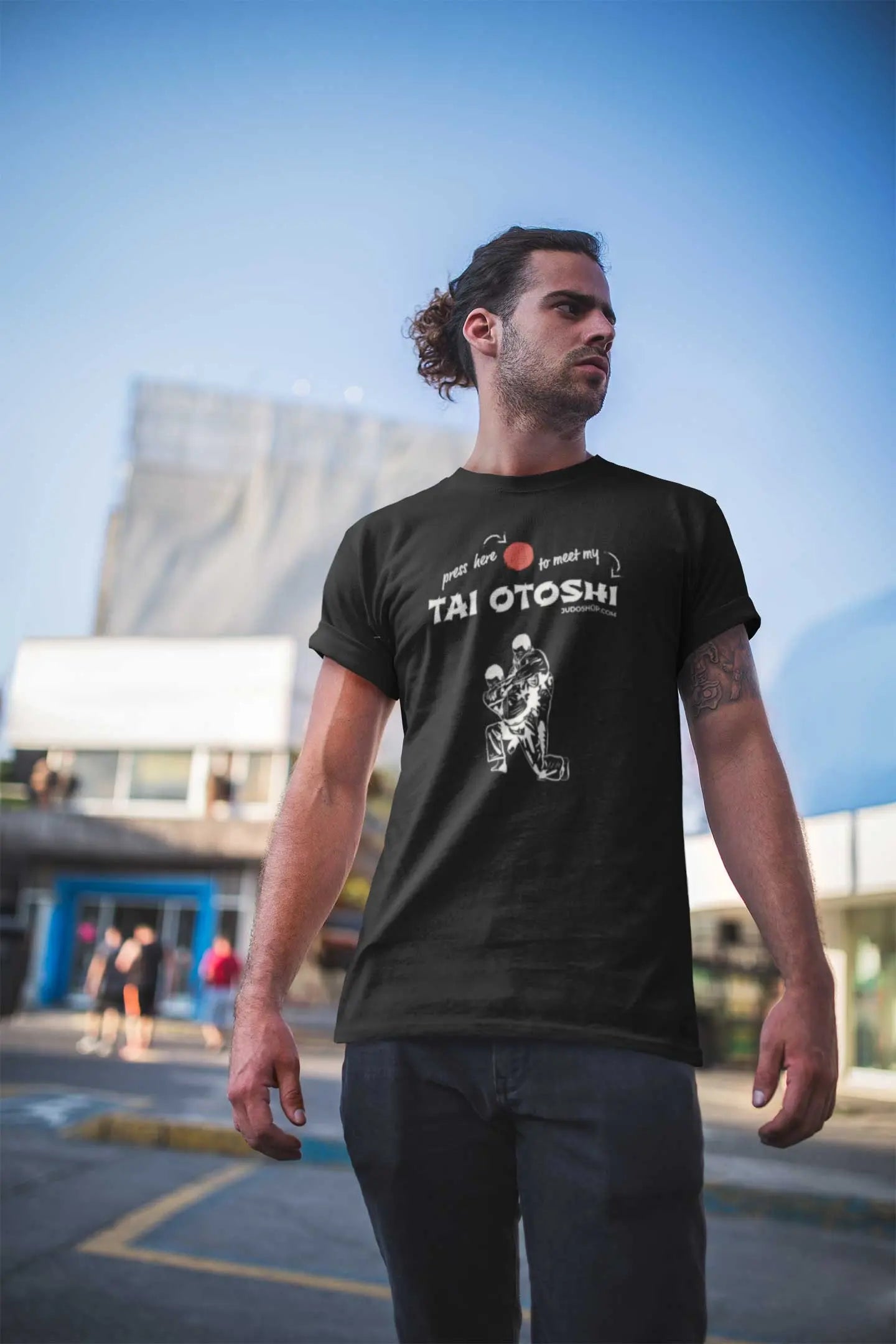 Man standing outdoors in a black 'Tai Otoshi Press Here' Judo T-Shirt, looking confidently into the distance.