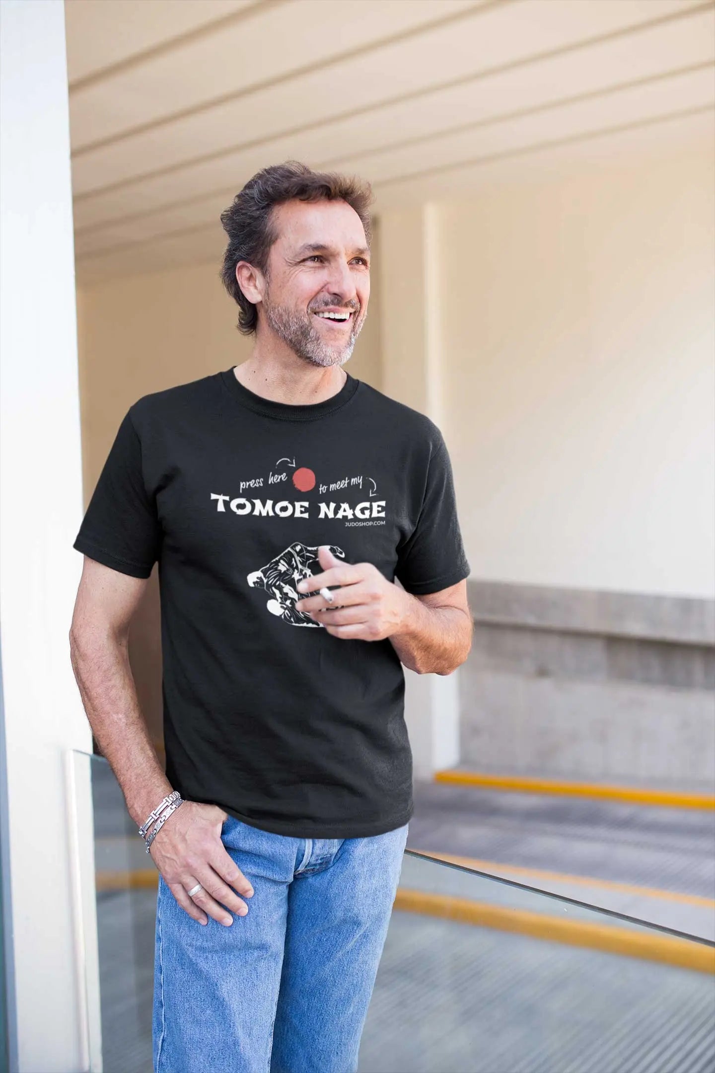 Man standing on a balcony wearing a black Judo T-Shirt with 'Press Here to Meet My Tomoe Nage' design, paired with blue jeans for a casual look.