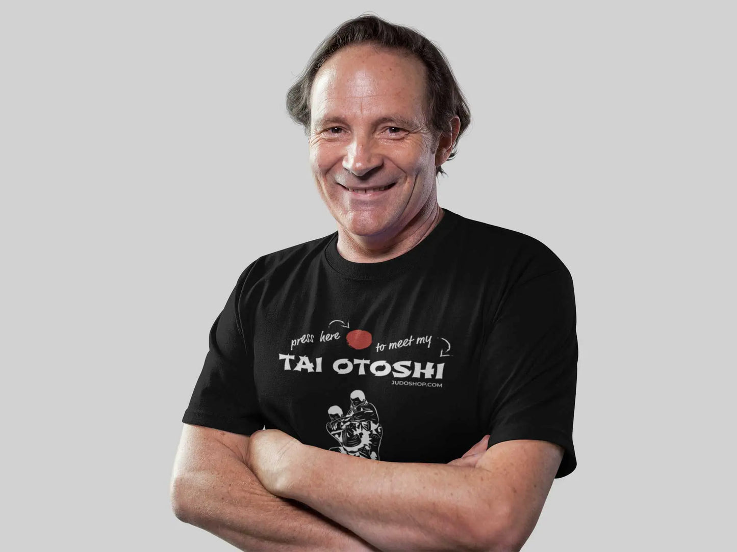 Smiling man crossing his arms while wearing a black 'Tai Otoshi Press Here' Judo T-Shirt, standing against a plain background.