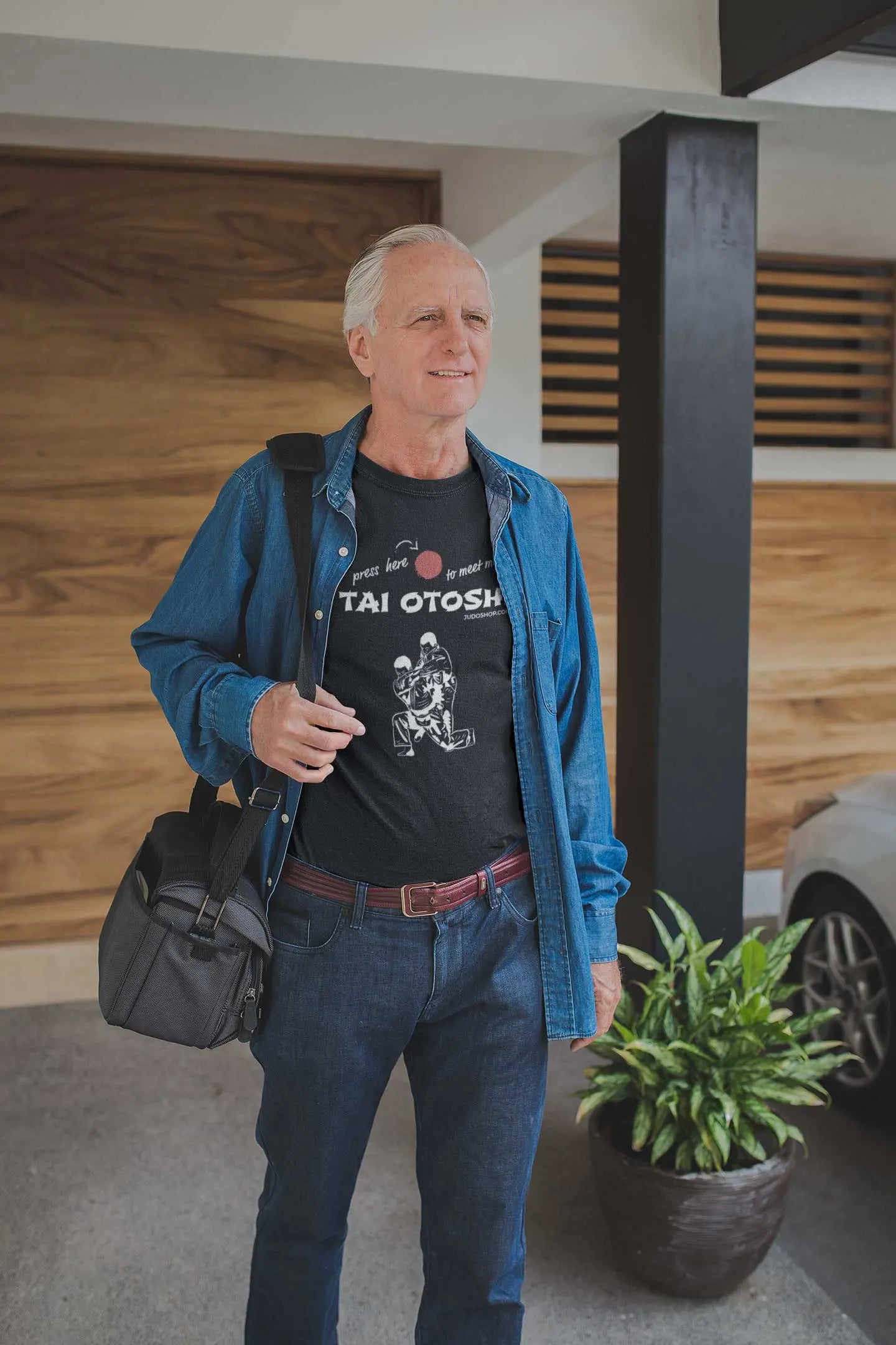 Man wearing a black 'Tai Otoshi Press Here' Judo T-Shirt and denim shirt, carrying a bag while standing near a car.
