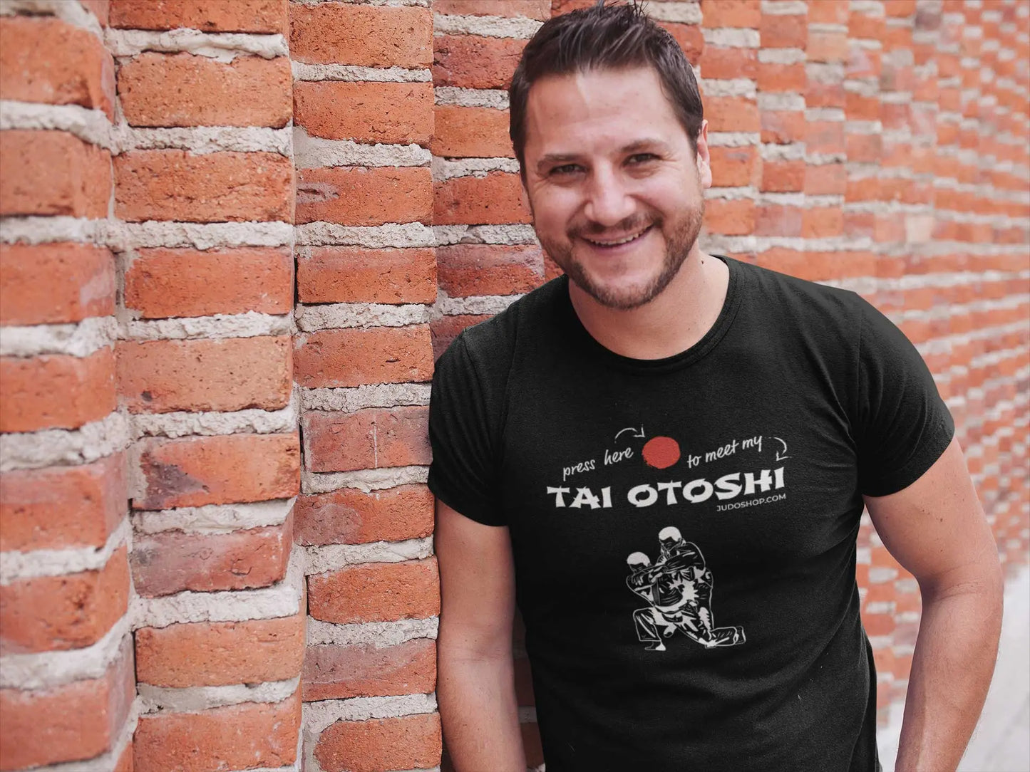 Man smiling and posing in a black 'Tai Otoshi Press Here' Judo T-Shirt, standing against a red brick wall.