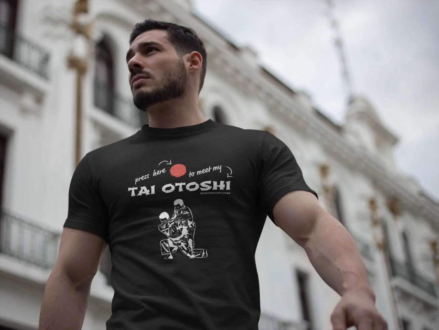 Man wearing a black 'Tai Otoshi Press Here' Judo T-Shirt, standing outdoors in front of a white building with a confident expression.