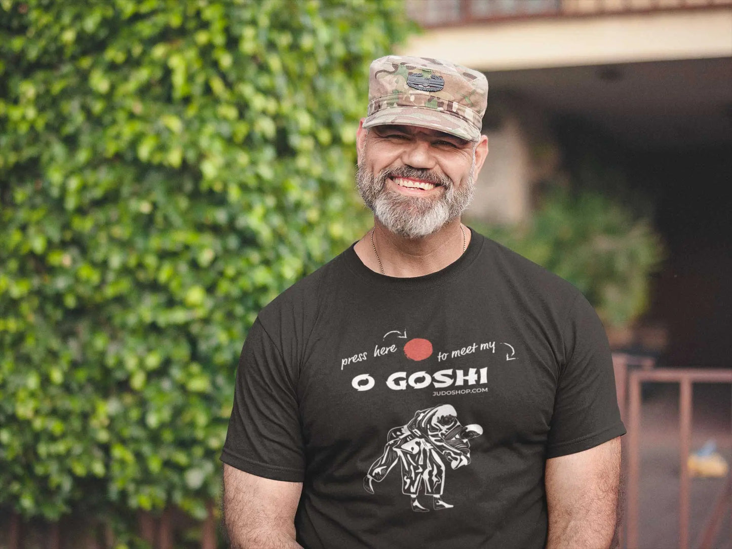 Man smiling outdoors in a black Judo T-Shirt with 'Press Here to Meet My O Goshi' design, styled with a camouflage cap for a rugged look.