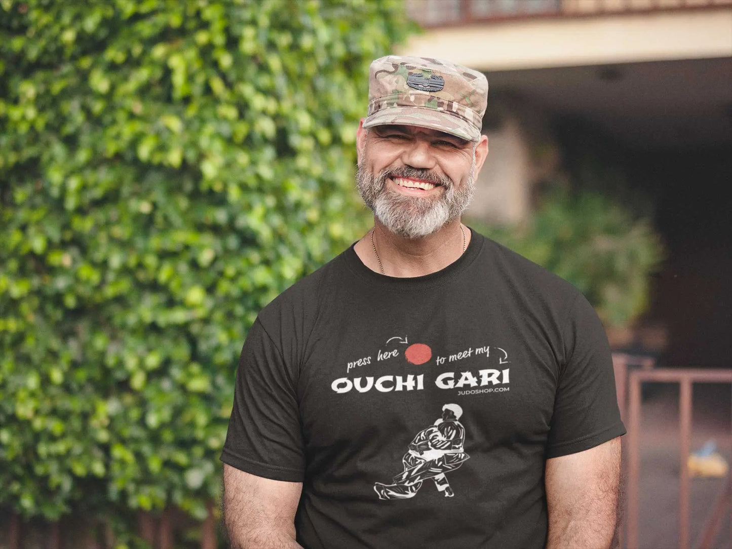 Man smiling outdoors in a black 'Ouchi Gari Press Here' Judo T-Shirt, styled with a camo cap for a rugged and sporty look.