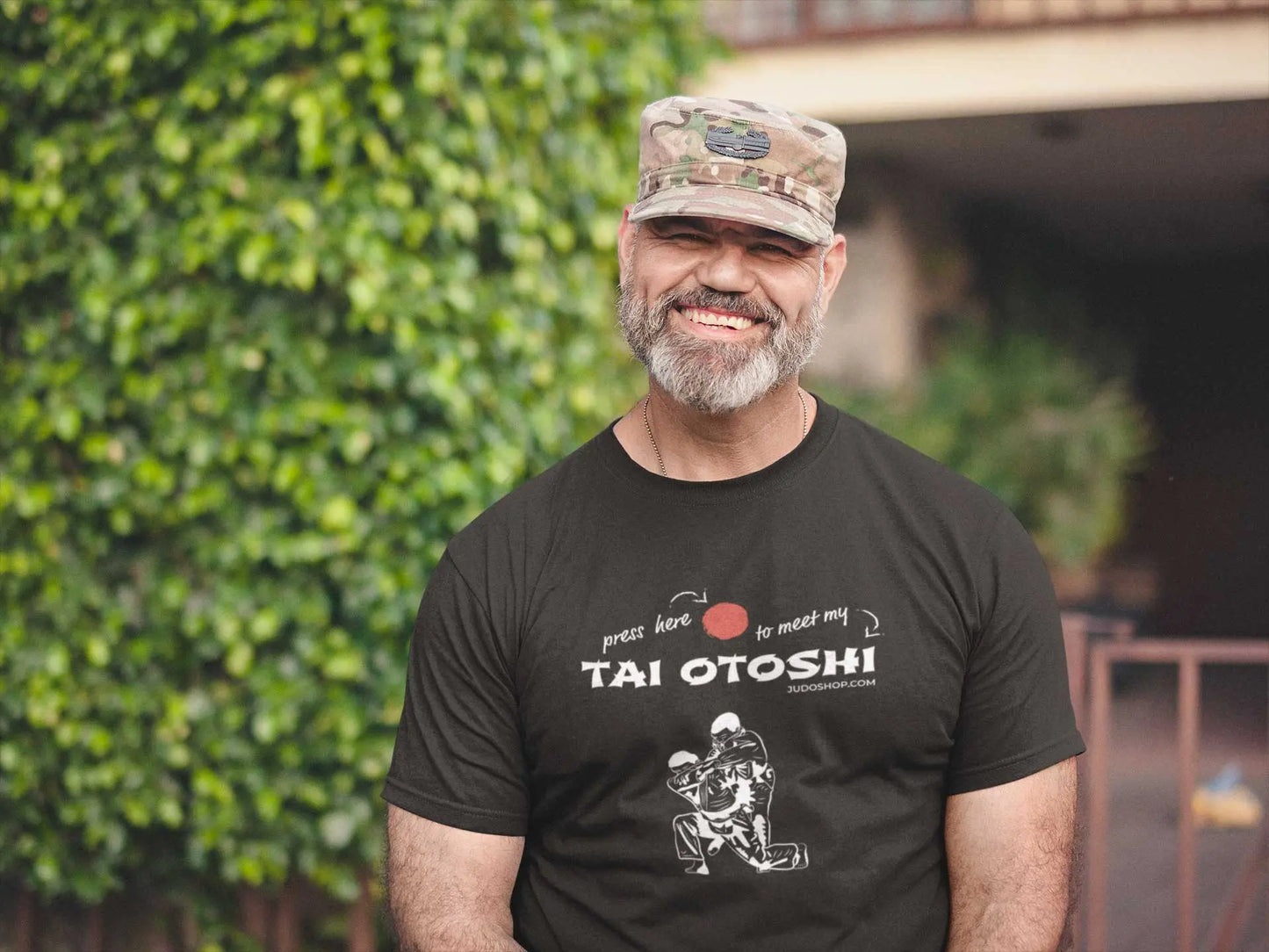 Man smiling and wearing a black 'Tai Otoshi Press Here' Judo T-Shirt, paired with a camouflage hat, standing outdoors.