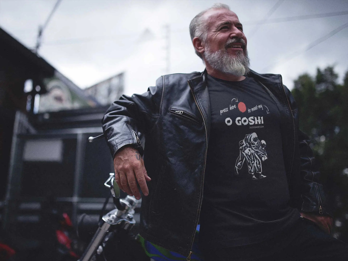 Man in a leather jacket posing with a motorcycle, wearing a black Judo T-Shirt featuring 'Press Here to Meet My O Goshi' design.