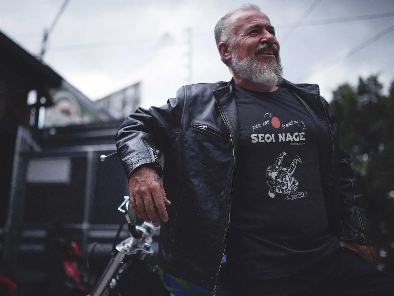 Bearded man in a black Seoi Nage Judo T-Shirt and leather jacket, leaning on a motorcycle in an urban setting.