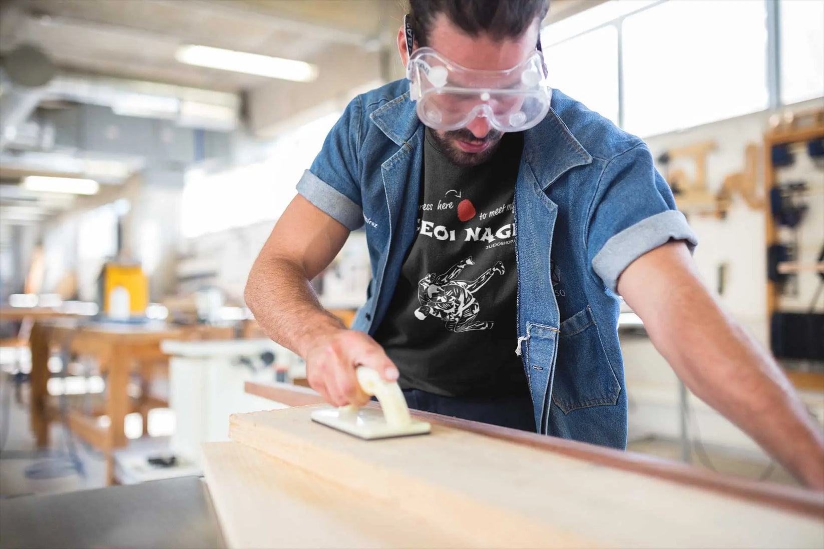 Man sanding wood in a workshop while wearing a black Seoi Nage Judo T-Shirt and protective goggles.