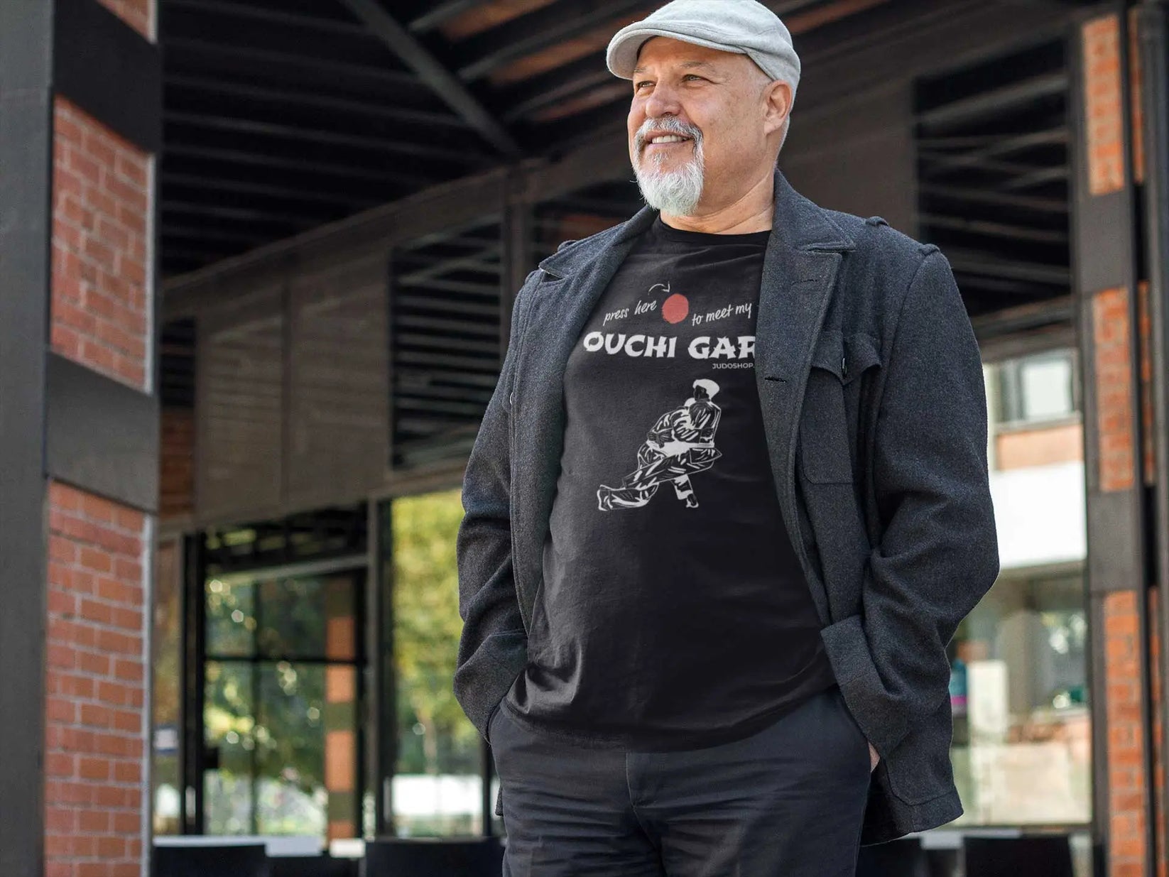 Man outdoors wearing a black 'Ouchi Gari Press Here' Judo T-Shirt, styled with a gray jacket for a casual look.