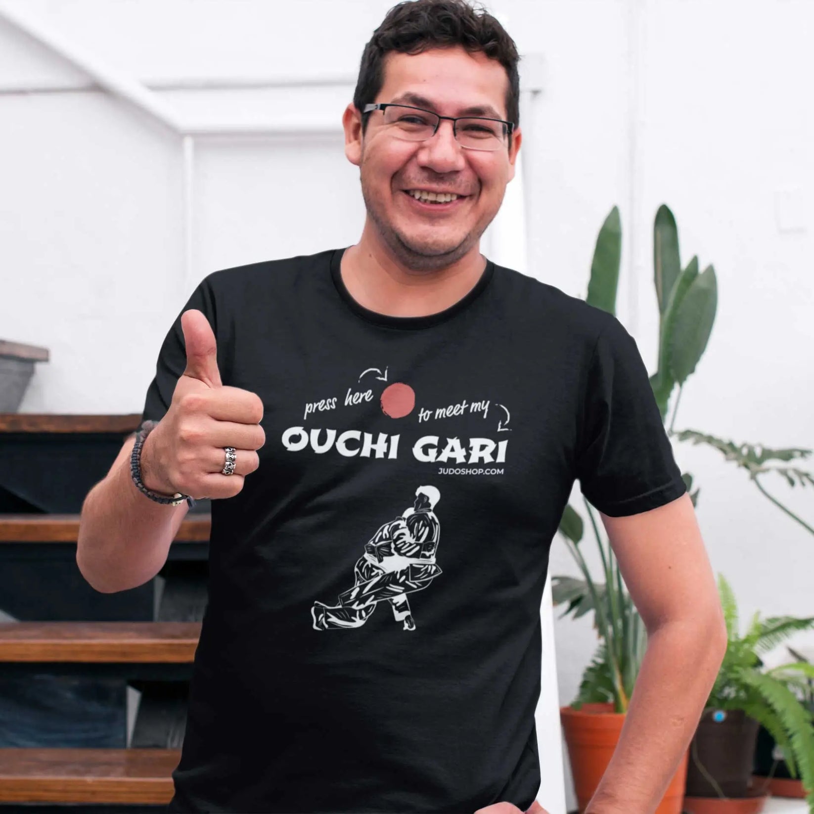 Smiling man giving a thumbs-up while wearing a black 'Ouchi Gari Press Here' Judo T-Shirt, showcasing a fun and sporty vibe.