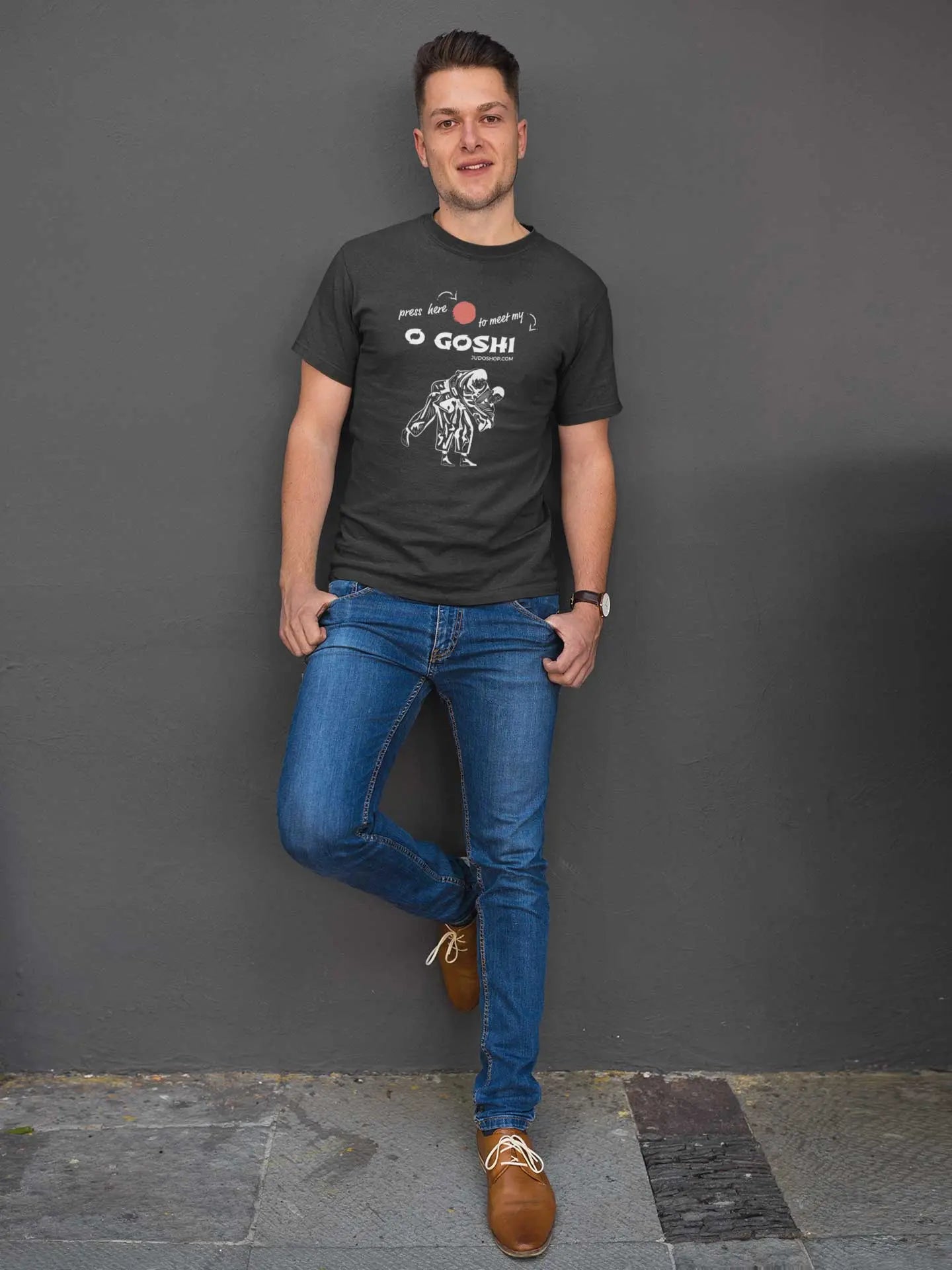 Man leaning against a wall in a black Judo T-Shirt with 'Press Here to Meet My O Goshi' design, paired with jeans for a relaxed style.