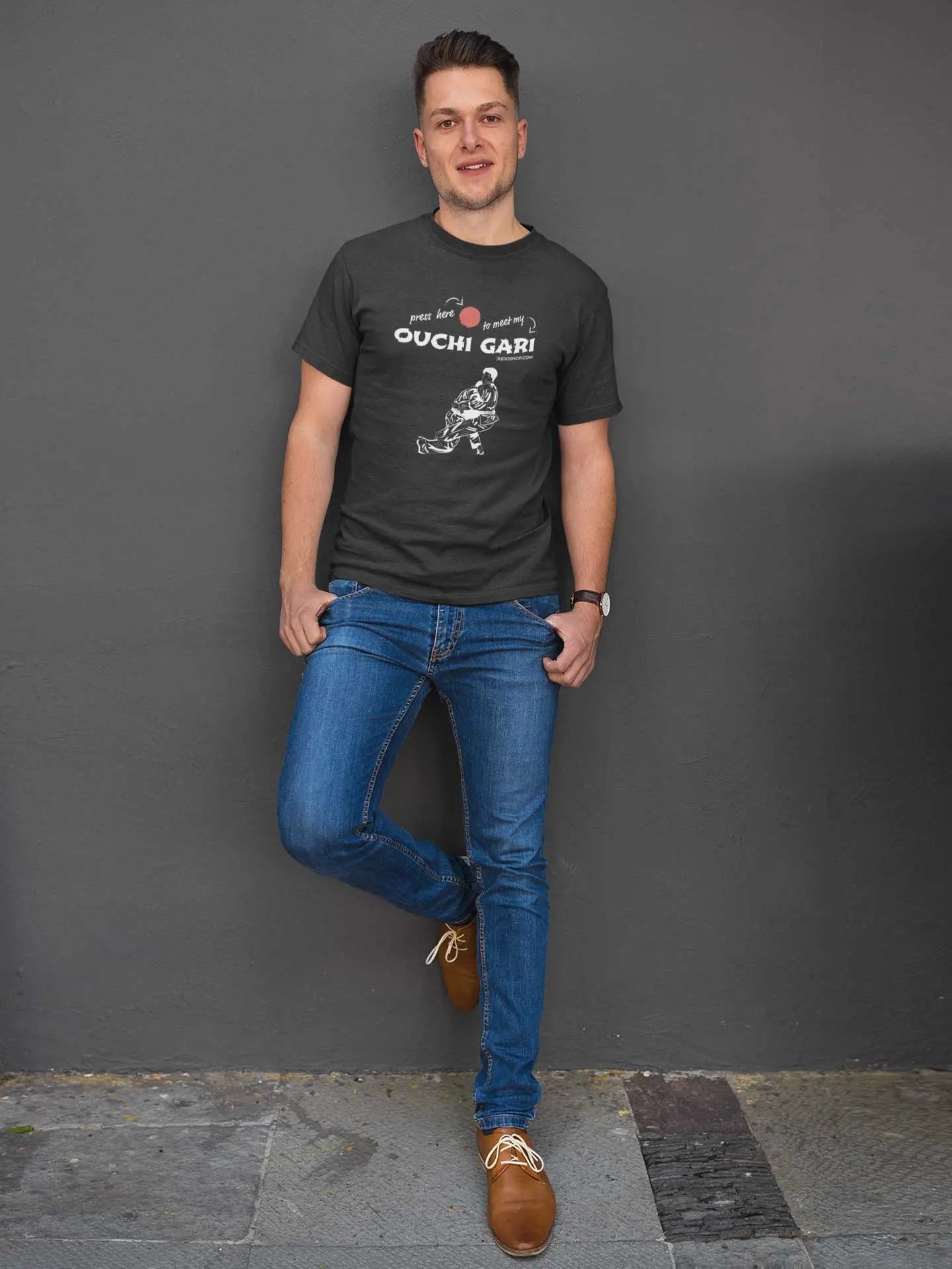 Man leaning against a wall in a black 'Ouchi Gari Press Here' Judo T-Shirt, paired with jeans for a relaxed and stylish look.