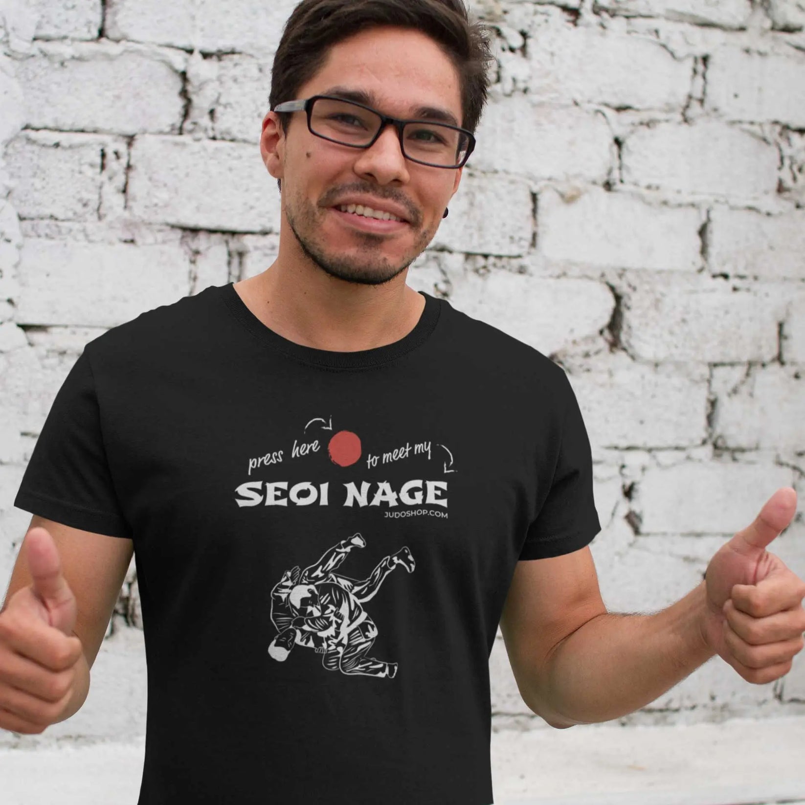 Man smiling and giving double thumbs-up while wearing a black Seoi Nage Judo T-Shirt, standing against a white brick wall.