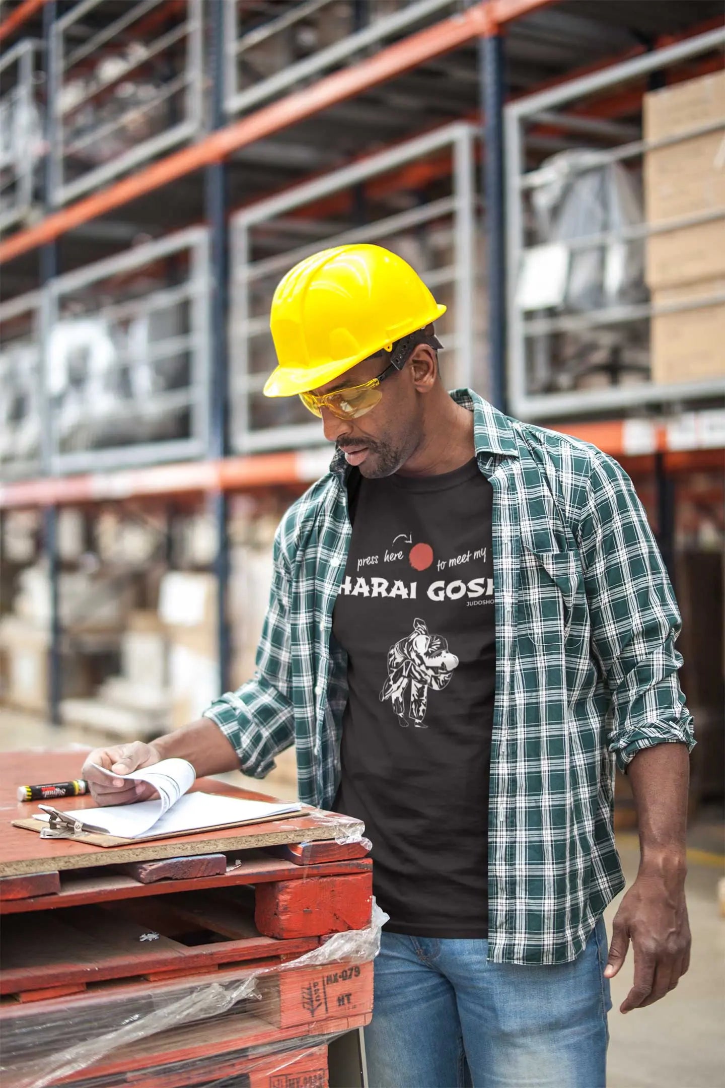 Man in a warehouse wearing a black 'Harai Goshi Press Here' Judo T-Shirt with a hard hat, blending workwear with casual fashion.