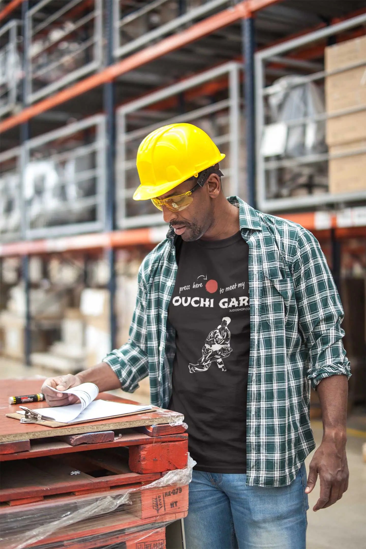 Man in a warehouse wearing a black 'Ouchi Gari Press Here' Judo T-Shirt, paired with a plaid shirt and hard hat for a rugged look.