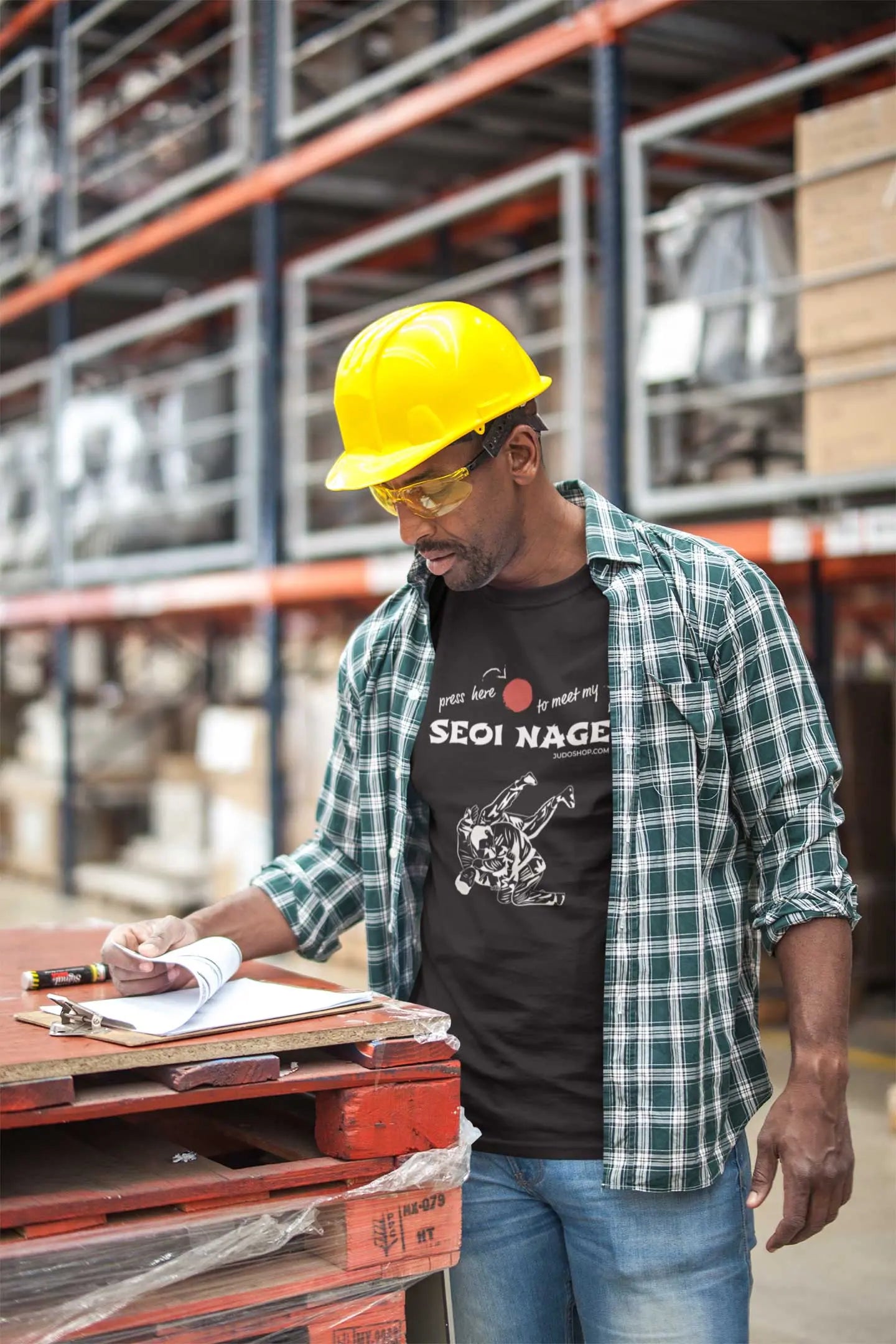 Man in a warehouse wearing a black Seoi Nage Judo T-Shirt, plaid shirt, and yellow hard hat, reviewing documents.