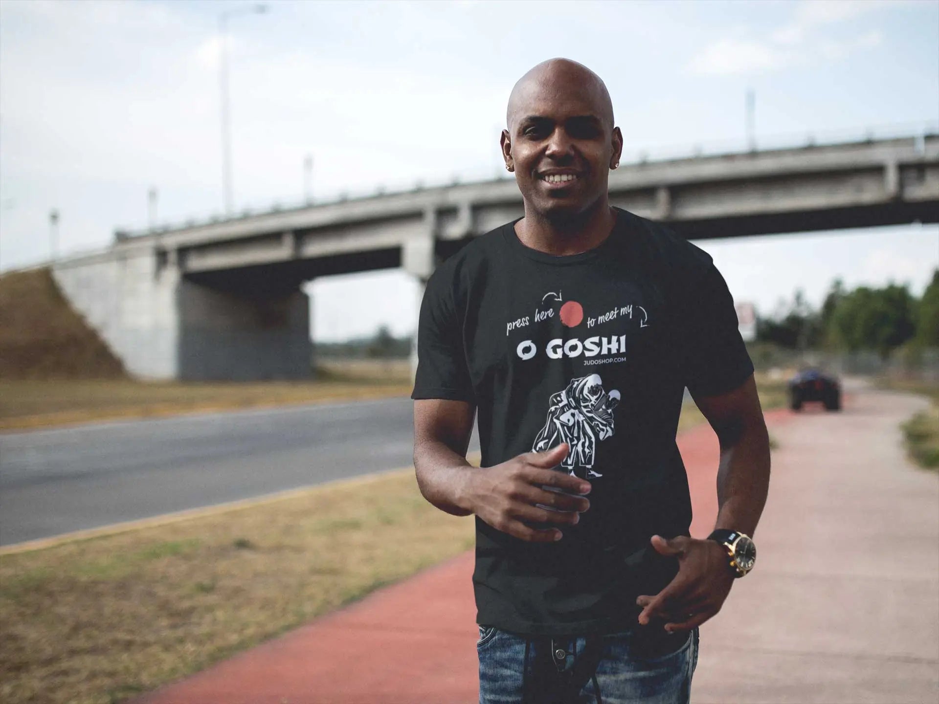 Man smiling outdoors in a black Judo T-Shirt featuring 'Press Here to Meet My O Goshi' text, perfect for casual and active wear.