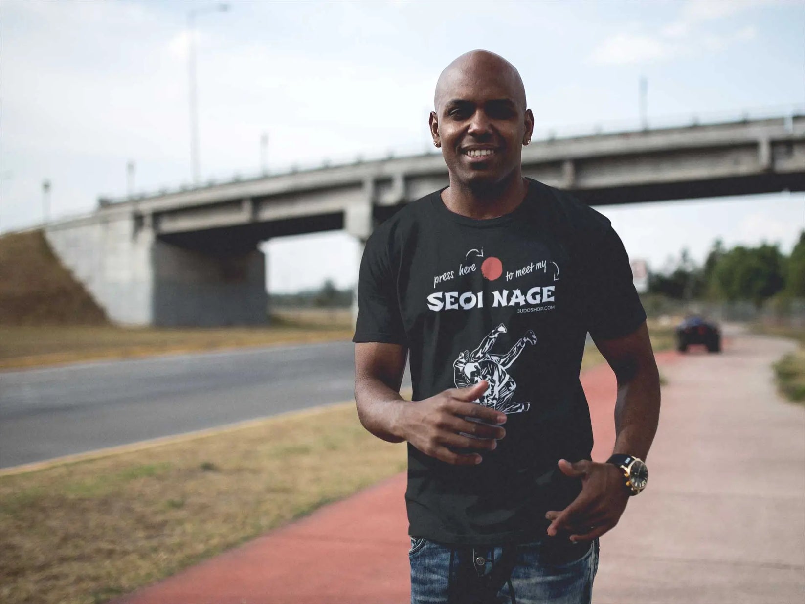 Man walking outdoors near a bridge, smiling while wearing a black Seoi Nage Judo T-Shirt.