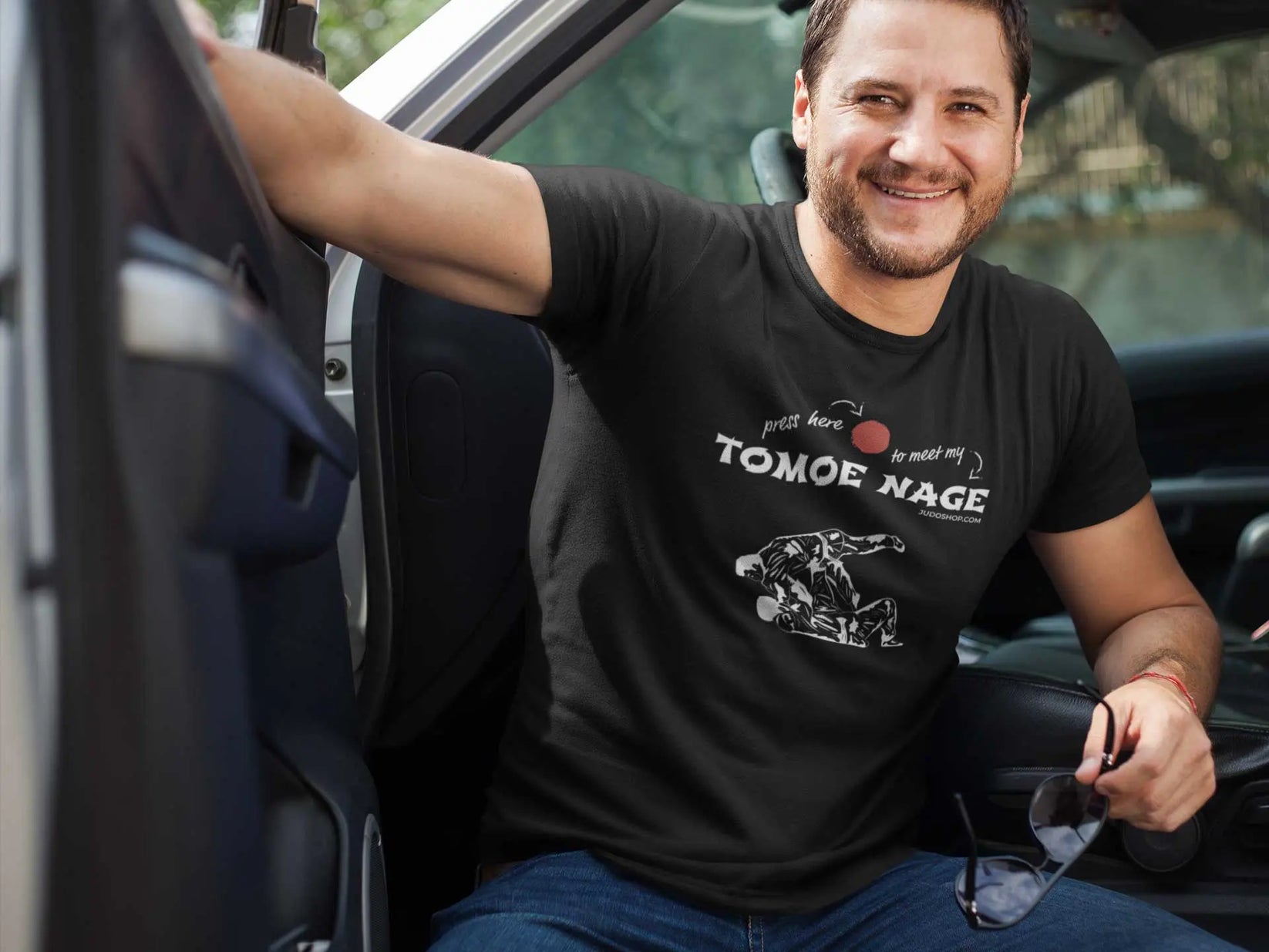 Man sitting in a car wearing a black Judo T-Shirt with 'Press Here to Meet My Tomoe Nage' design, styled for a casual outing.