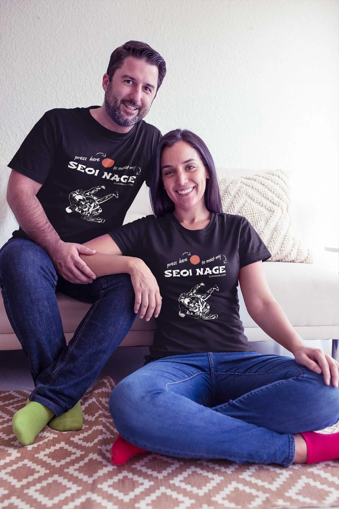 Couple sitting on a couch, both wearing black Seoi Nage Judo T-Shirts, smiling and posing together.
