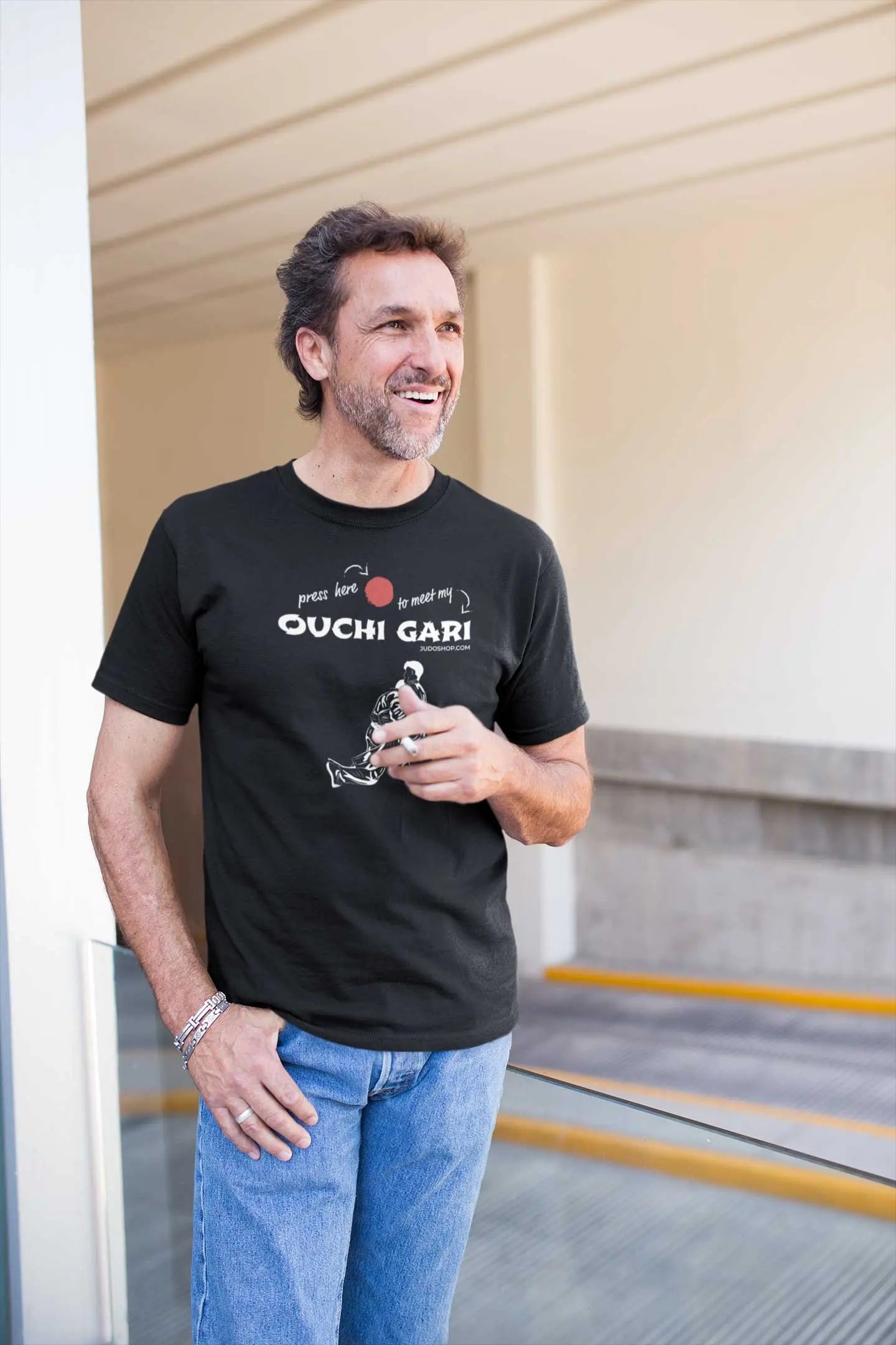Man standing on a balcony wearing a black 'Ouchi Gari Press Here' Judo T-Shirt, styled with jeans for a casual outfit.