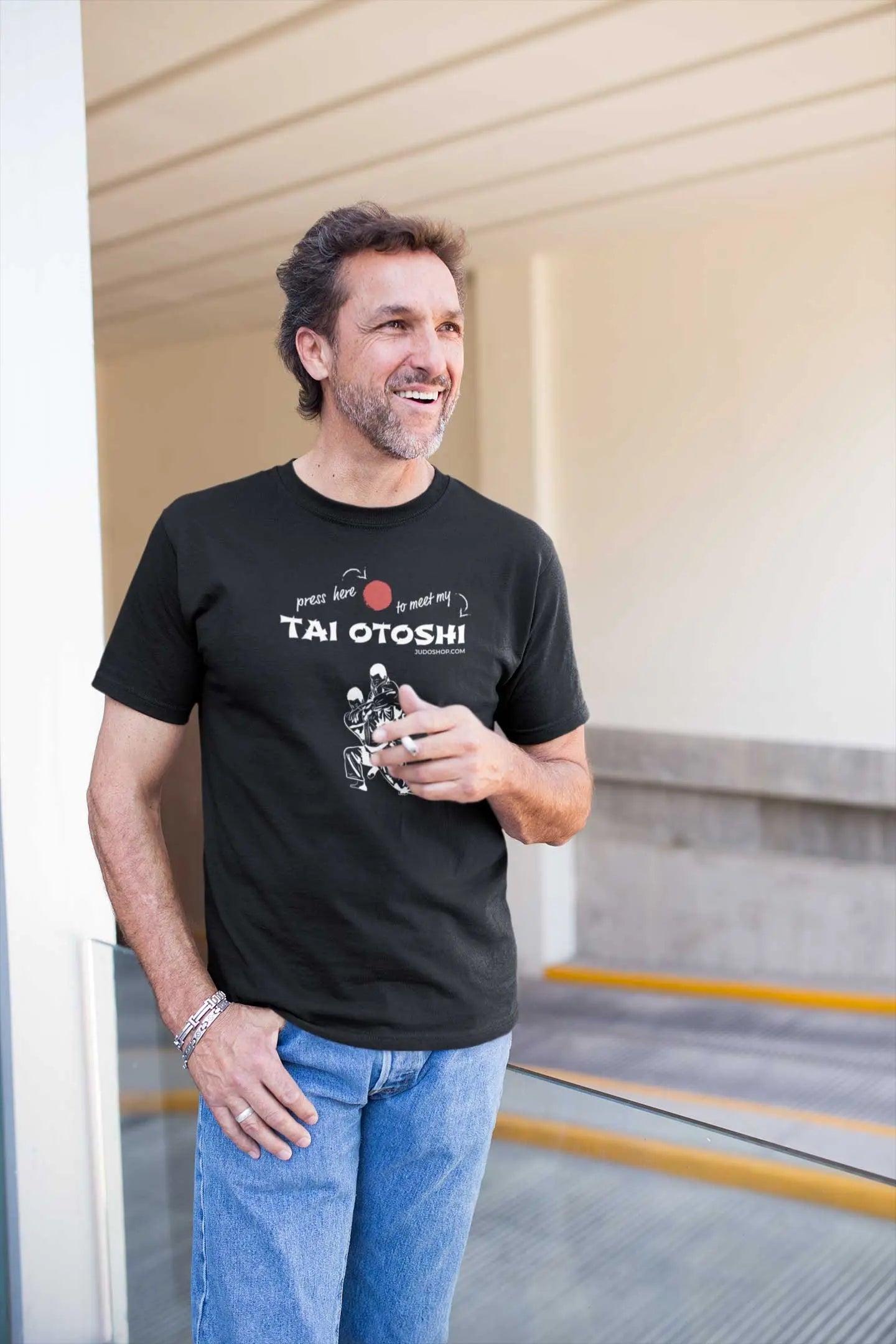 Man wearing a black 'Tai Otoshi Press Here' Judo T-Shirt, standing on a balcony and smiling while holding a cigarette.