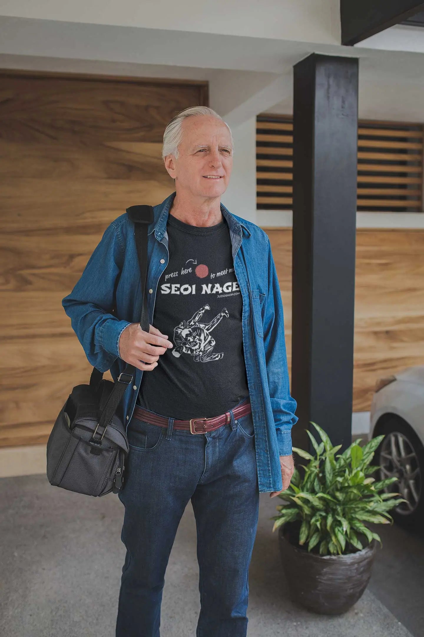 Man wearing a black Seoi Nage Judo T-Shirt and denim shirt, carrying a bag while standing in a modern interior.