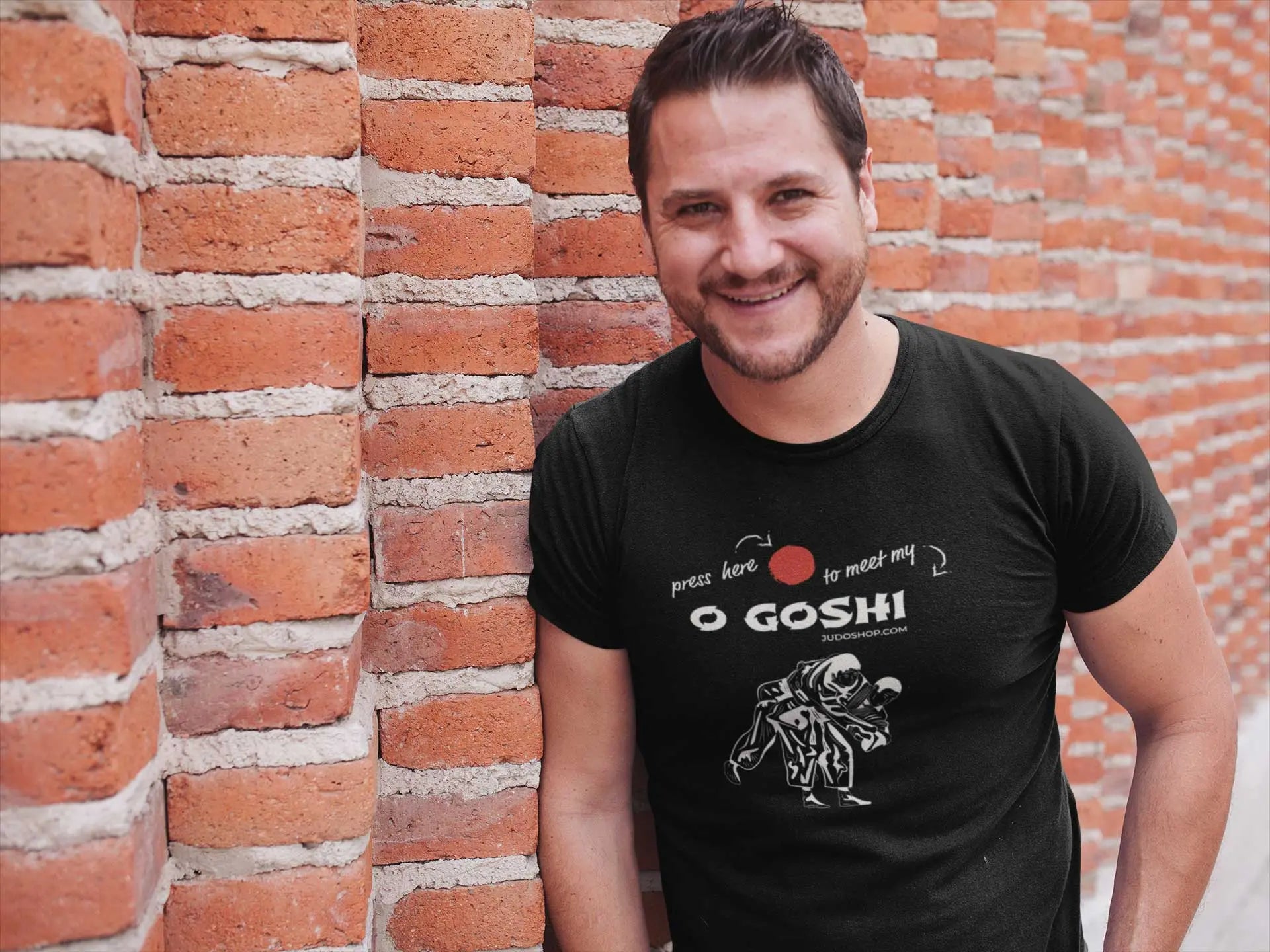 Man smiling against a brick wall, wearing a black Judo T-Shirt featuring 'Press Here to Meet My O Goshi' text and martial arts graphic.