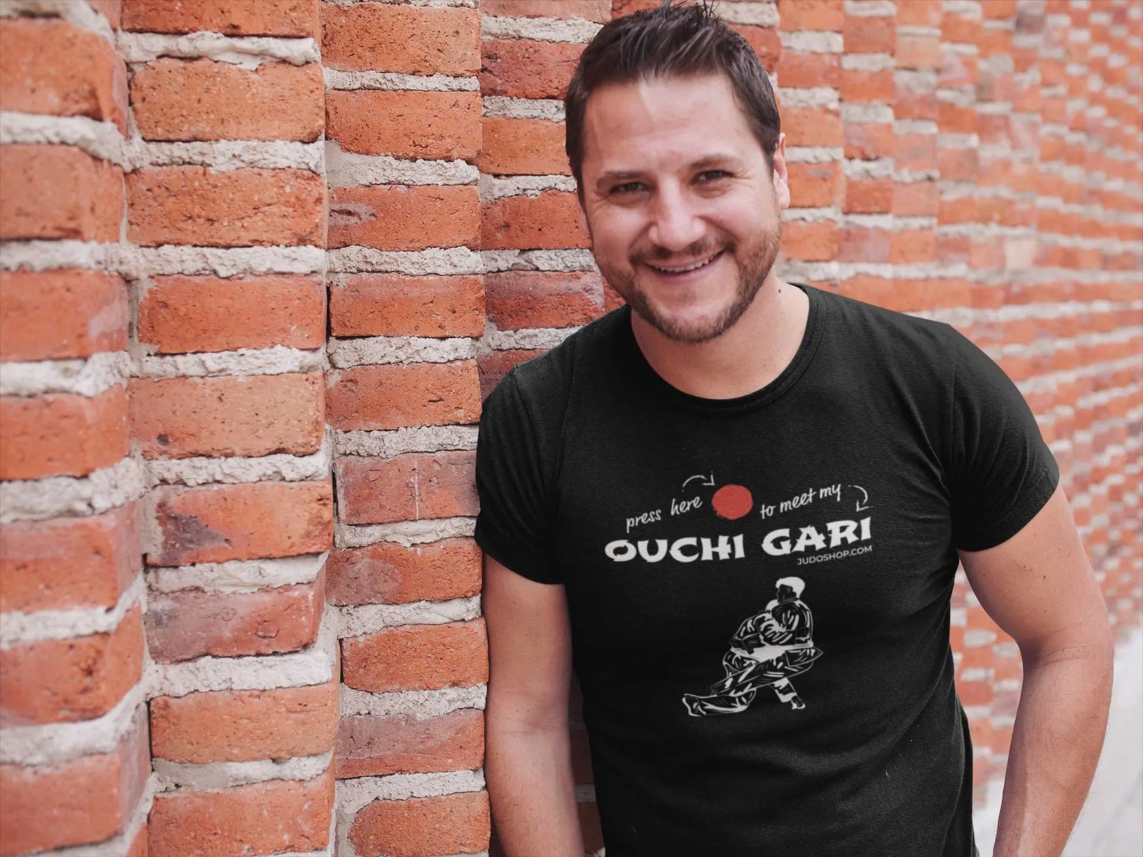 Man smiling against a brick wall wearing a black 'Ouchi Gari Press Here' Judo T-Shirt, perfect for casual and sporty occasions.