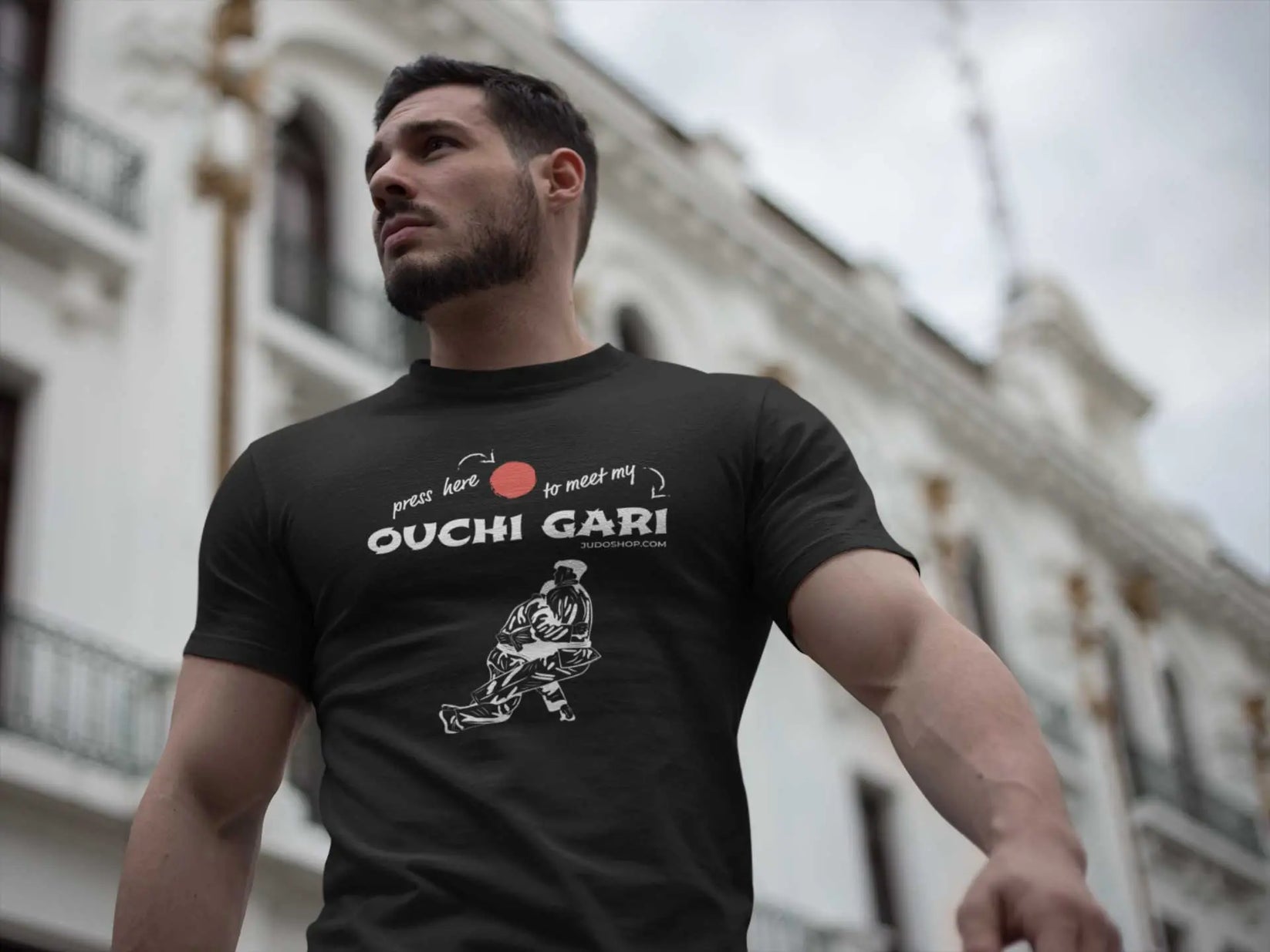Man walking in an urban setting wearing a black 'Ouchi Gari Press Here' Judo T-Shirt, ideal for casual and athletic wear.