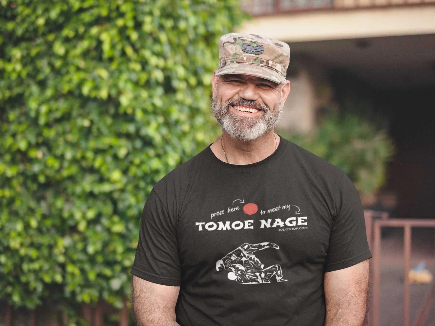 Man in a camouflage cap wearing a black Judo T-Shirt with 'Press Here to Meet My Tomoe Nage' design, styled for a casual outdoor look.