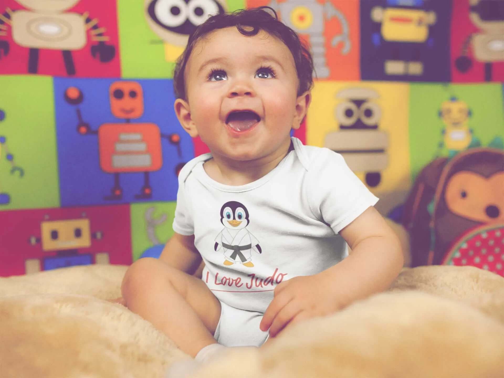 Adorable baby wearing a white Judo Baby Bodysuit featuring a cute penguin design with 'I Love Judo' text, sitting on a soft beige surface with a colorful robot-themed background.