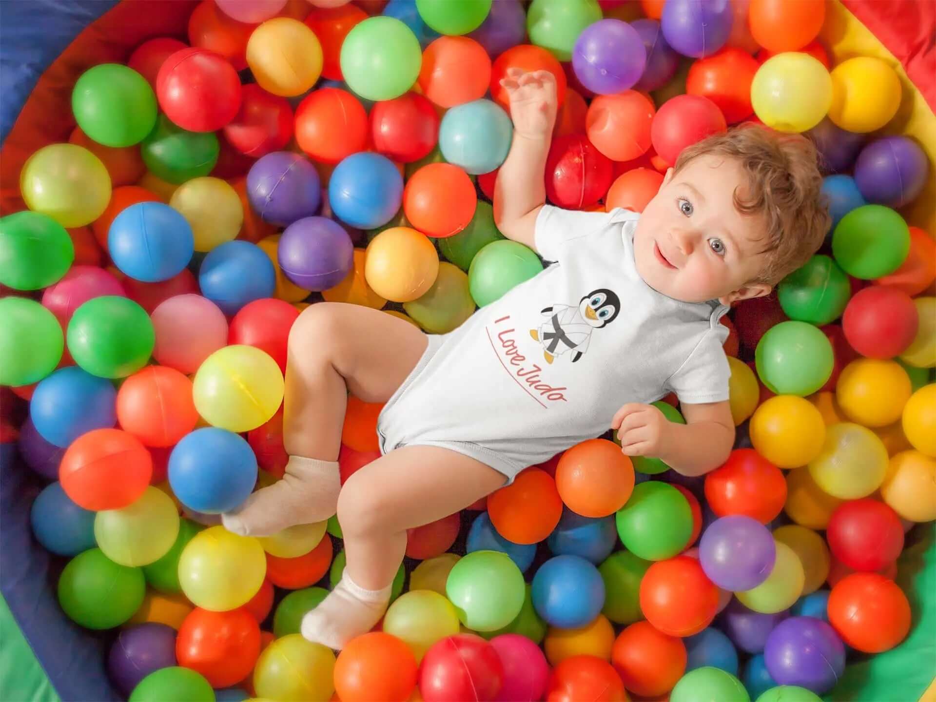 Baby wearing a white Judo bodysuit with a penguin and 'I Love Judo' design, lying in a colorful ball pit. Perfect for toddlers.