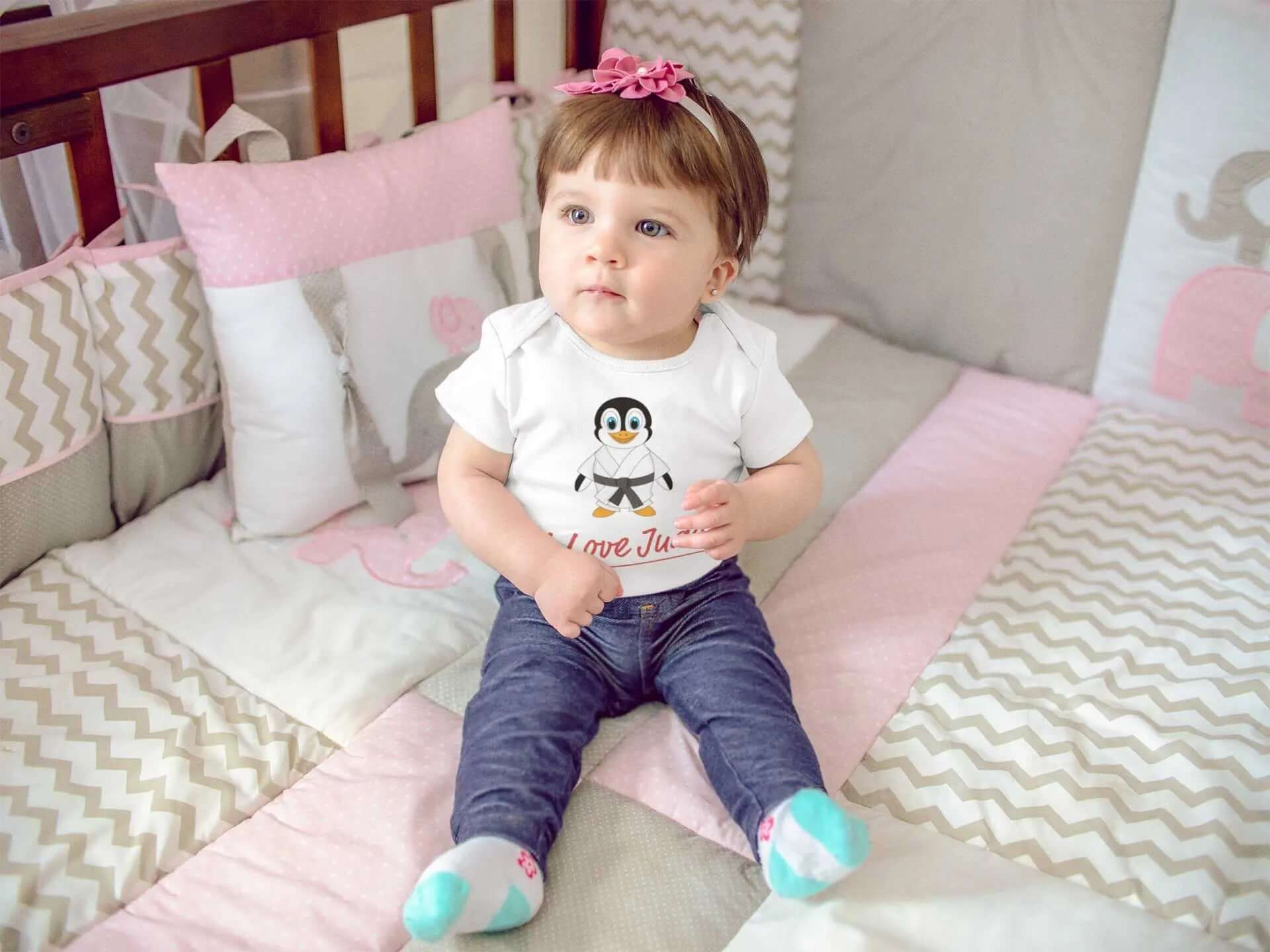Baby wearing a white Judo bodysuit featuring a cute penguin design and 'I Love Judo' text, sitting in a crib with pastel bedding.