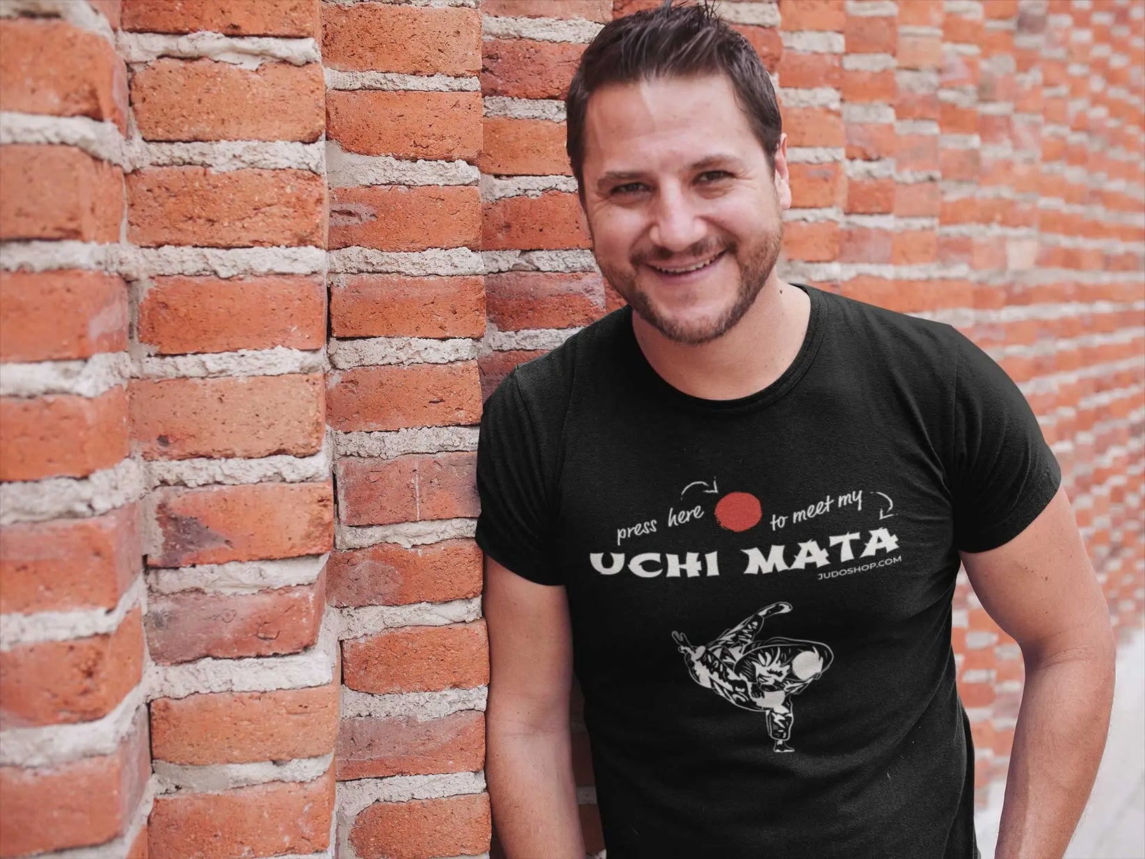 Man leaning against a brick wall wearing a black Judo T-Shirt with 'Press here to meet my Uchi Mata' design, casual and trendy.